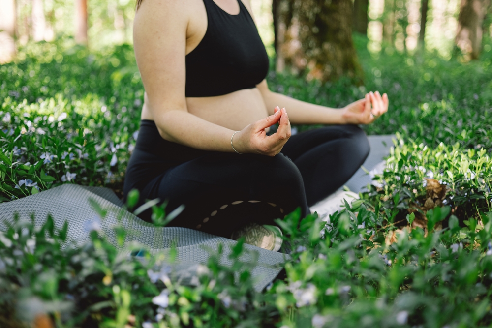 Expectant mother sitting cross-legged on a yoga mat in a green outdoor setting, focusing on relaxation, wellness, and holistic prenatal care – trusted women’s health center