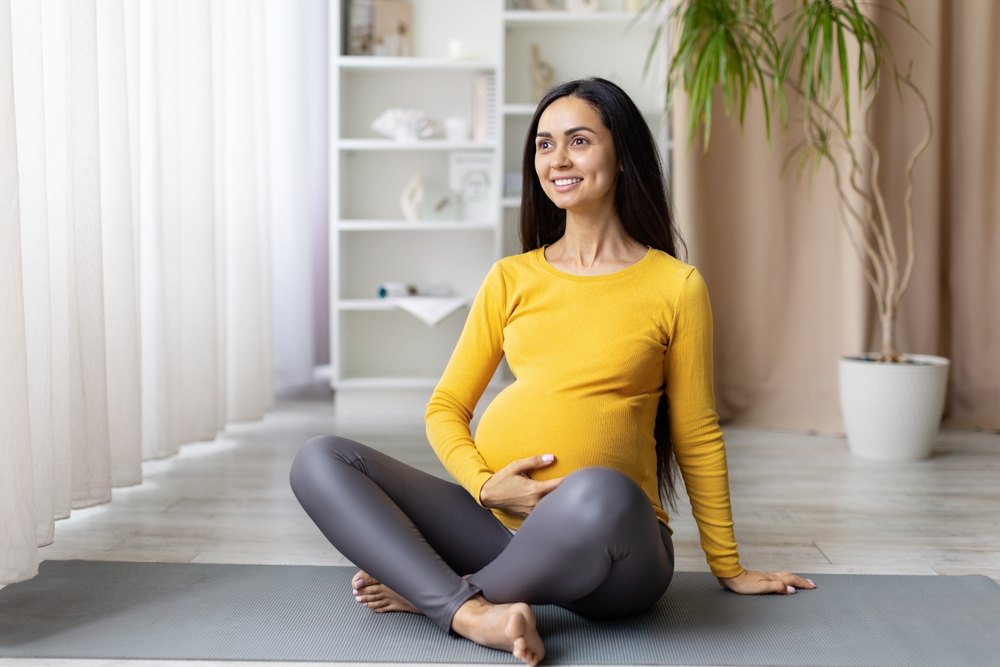 Smiling pregnant woman sitting cross legged on yoga mat, gently holding belly