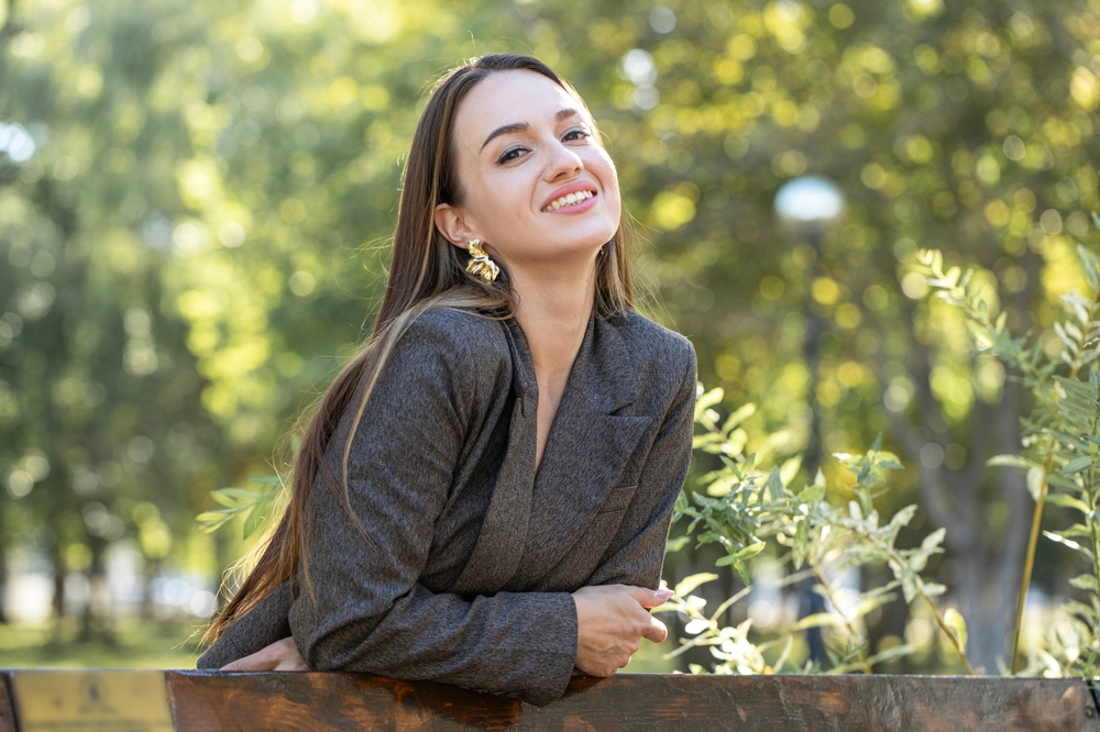 Professional woman smiling while leaning on a wooden railing in a green outdoor space, symbolizing confidence, wellness, and compassionate care – trusted women’s health center
