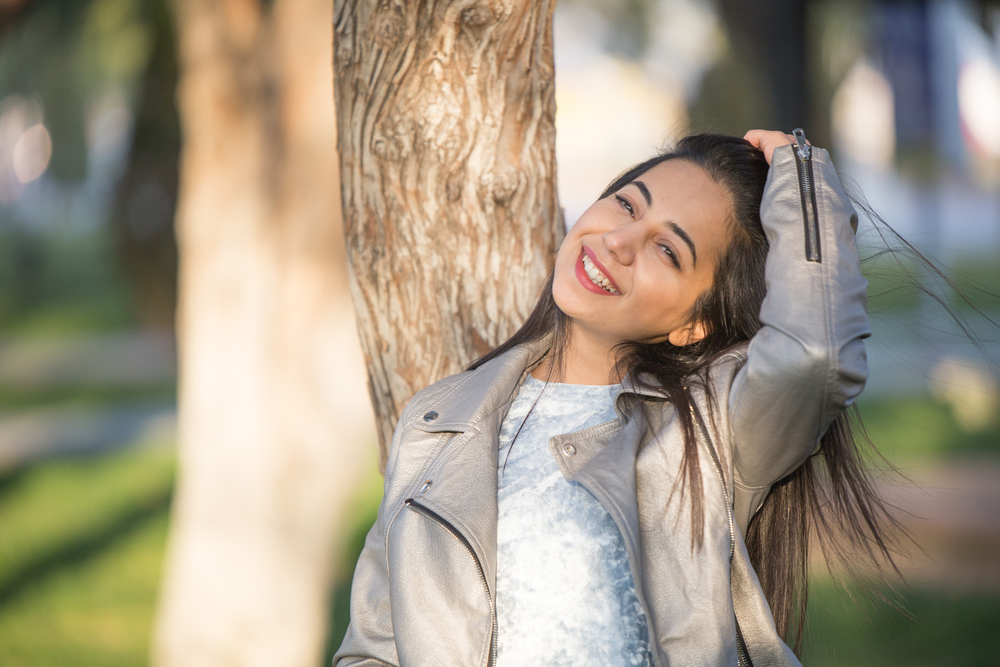 Relaxed woman smiling while leaning against a tree in a sunlit park, representing comfort, self-care, and supportive wellness services – trusted women’s health center