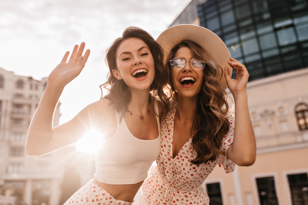 Two happy women laughing and waving outdoors in a bright urban setting, symbolizing friendship, confidence, and supportive women’s healthcare – obgyn margate