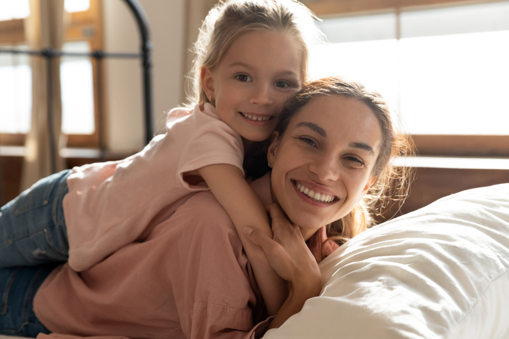 Smiling mother and young daughter embracing on a bed in a bright room, symbolizing family-centered care, trust, and compassionate women’s healthcare – obgyn north miami beach