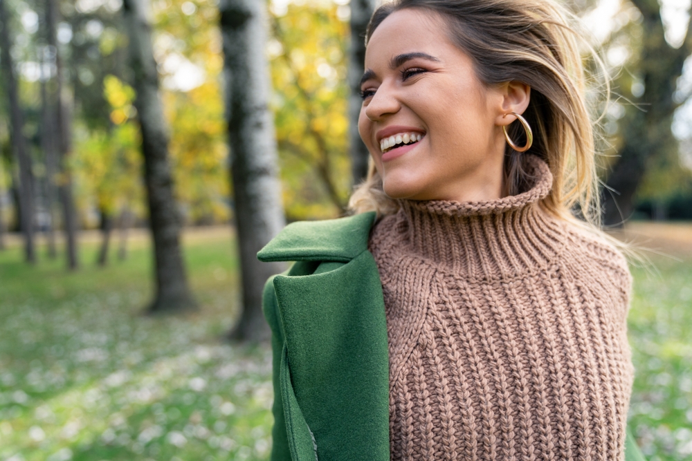 Smiling woman enjoying a peaceful outdoor moment representing comfort and wellness – gynecologist homestead fl Woman smiling while walking through a tree-lined park in a cozy sweater, reflecting confidence, relaxation, and compassionate women’s healthcare – gynecologist homestead fl